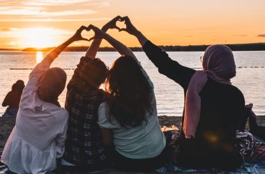 four people sitting on shore forming hearts with their hands during golden hour