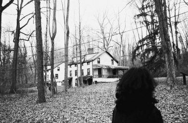 grayscale photo of woman in black coat standing near bare trees