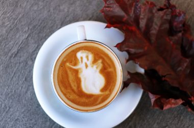 white ceramic teacup filled with ghost illustration coffee latte on white ceramic saucer beside maroon leaf photography