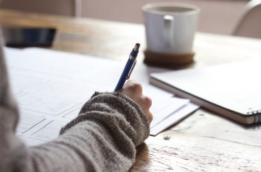 person writing on brown wooden table near white ceramic mug