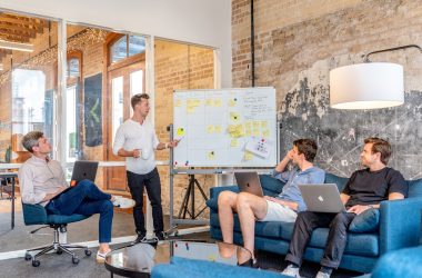 three men sitting while using laptops and watching man beside whiteboard