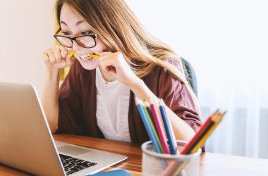 woman biting pencil while sitting on chair in front of computer during daytime