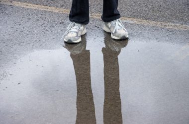 A person standing in the middle of a street with their reflection in a puddle